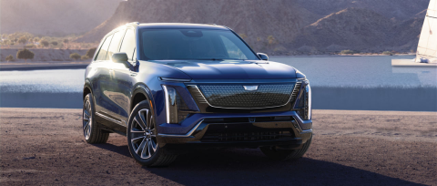 The image shows a dark blue 2026 Cadillac VISTIQ, an all-electric SUV, positioned on a dirt surface with a blurred background featuring a body of water and hills.