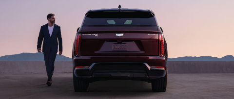 The image shows the rear view of a dark red 2025 Cadillac Escalade IQ, with a man in a suit walking toward the left side of the vehicle.